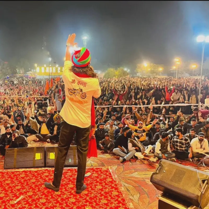 Man wearing an Om Banna logo shirt on stage with a large cheering crowd in the background